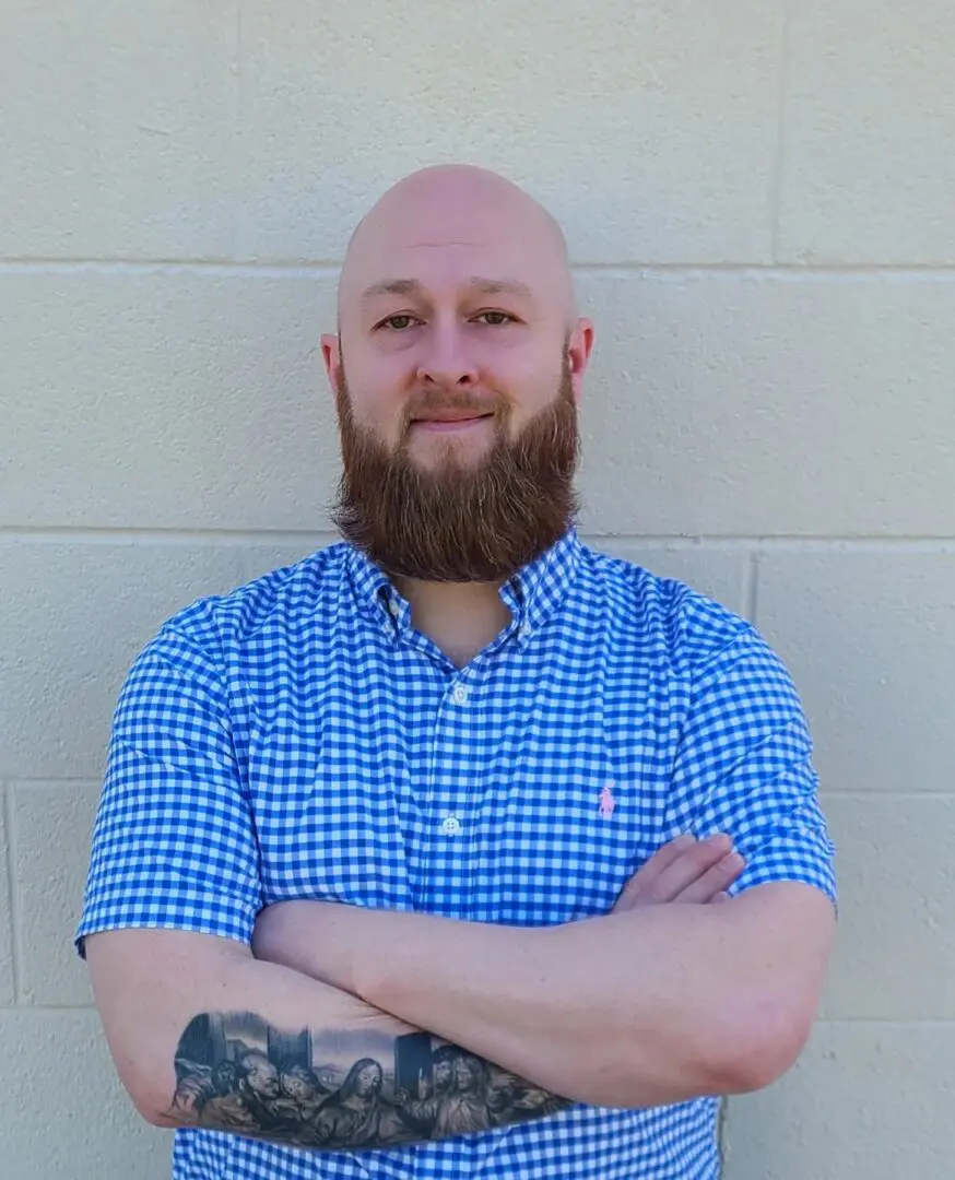 A man with beard and blue shirt standing in front of a wall.